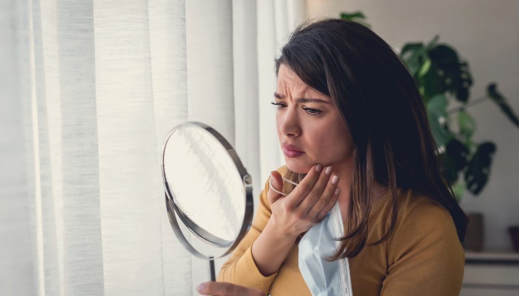 Woman looking herself in the mirror at home standing by the big window. She is concerned about acne, maskne