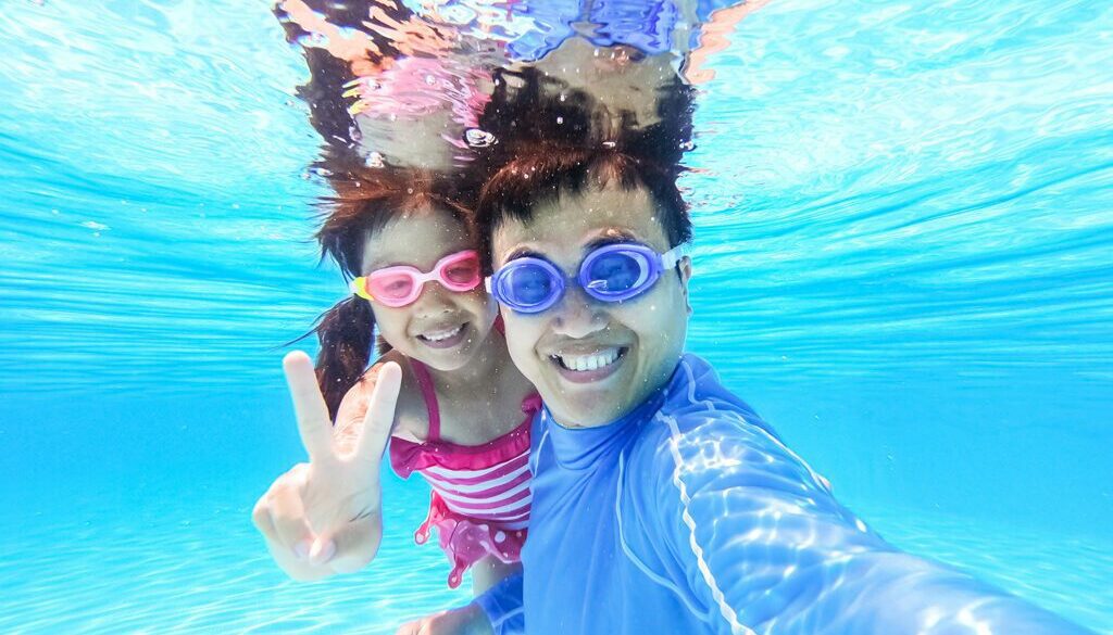 Asian dad and daughter smiling under water How do you get Swimmer's Ear?