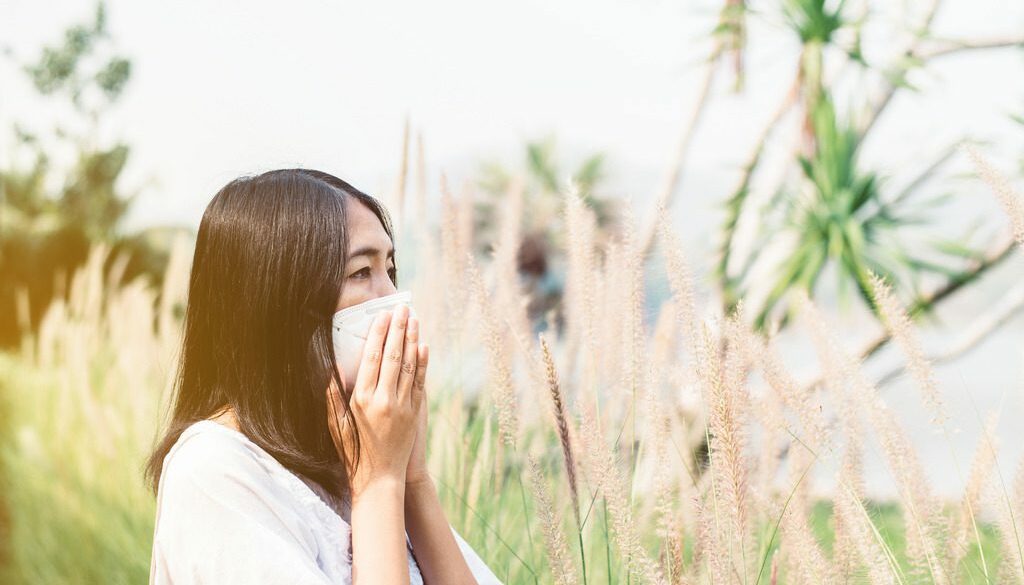 An Asian Woman Standing Amongst Weeds Covering Her Nose Dealing with Ragweed Allergy Symptoms.