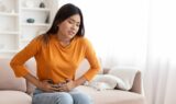 An Asian woman sitting on the couch holding her lower stomach in pain living with endometriosis