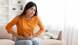 An Asian woman sitting on the couch holding her lower stomach in pain living with endometriosis